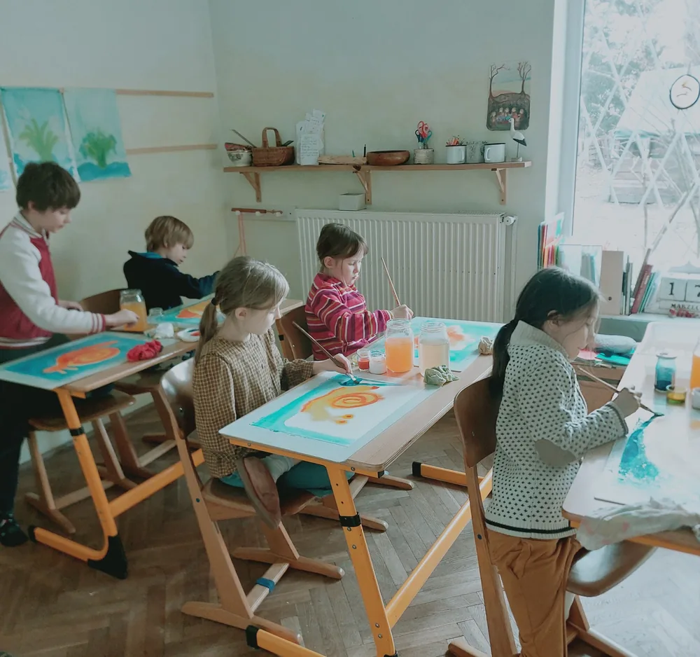 Czworo dzieci maluje akwarelami duże prace przy drewnianych stolikach w sali